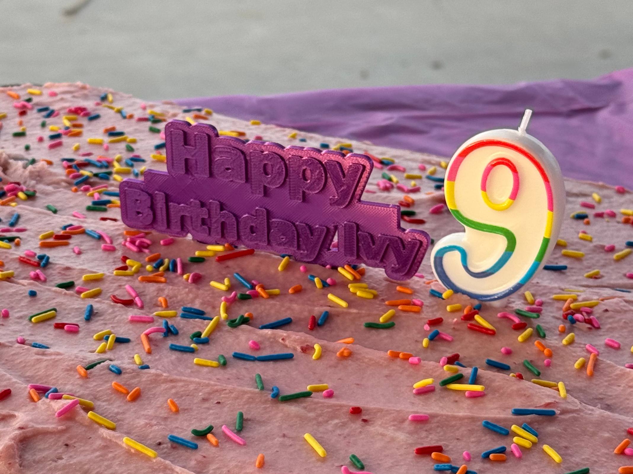 3D printed 'Happy Birthday Ivy' cake topper in purple silk PLA on a pink birthday cake with sprinkles